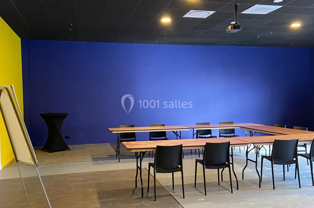 Salle de réunion avec murs colorés, tables en bois disposées en U, chaises noires et un tableau blanc sur pied.