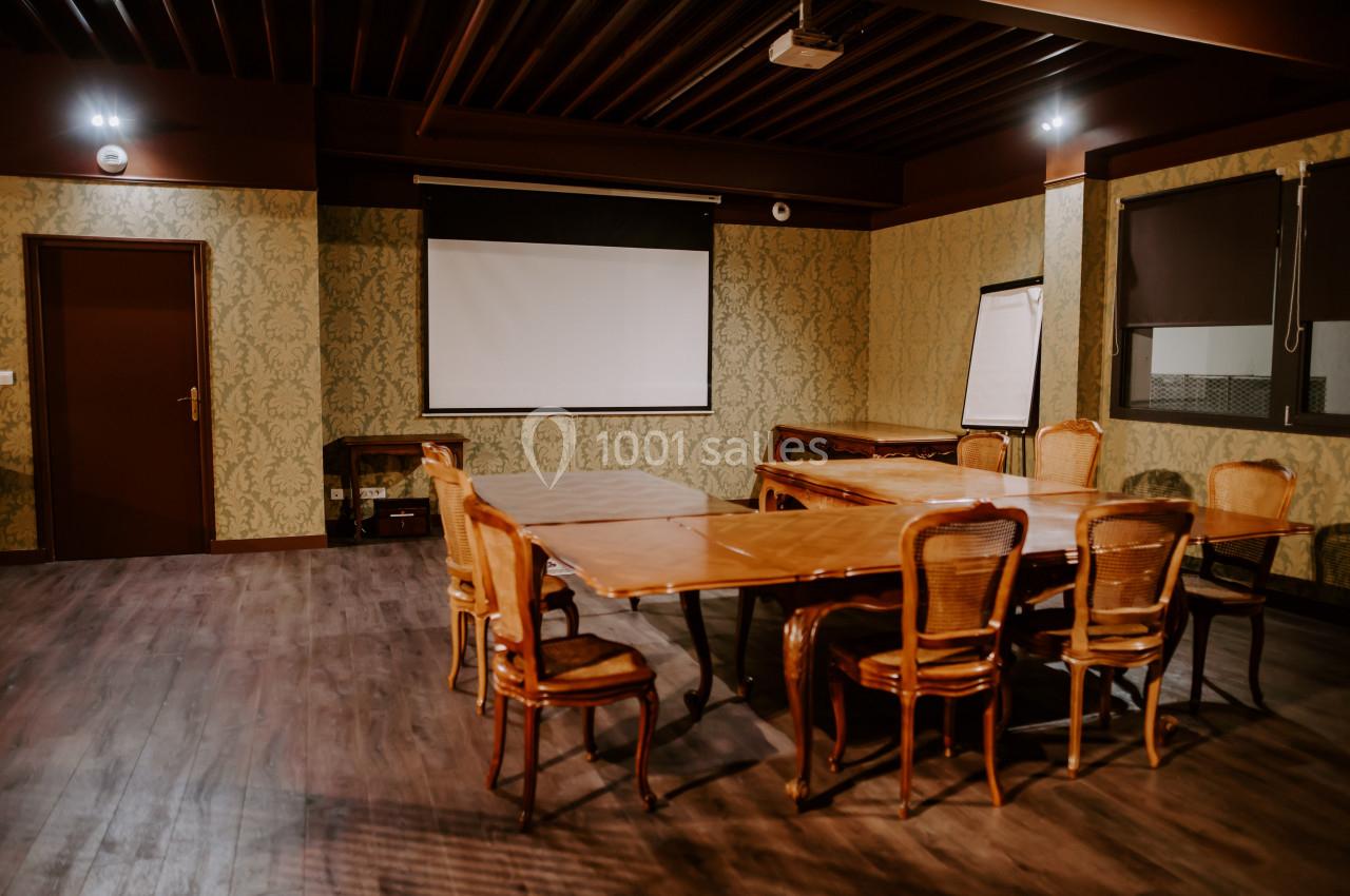 Salle de réunion avec table en bois, chaises en osier, écran de projection, paperboard et éclairage tamisé.