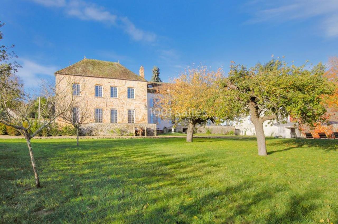 Grande maison en pierre entourée d'un jardin arboré sous un ciel bleu clair.
