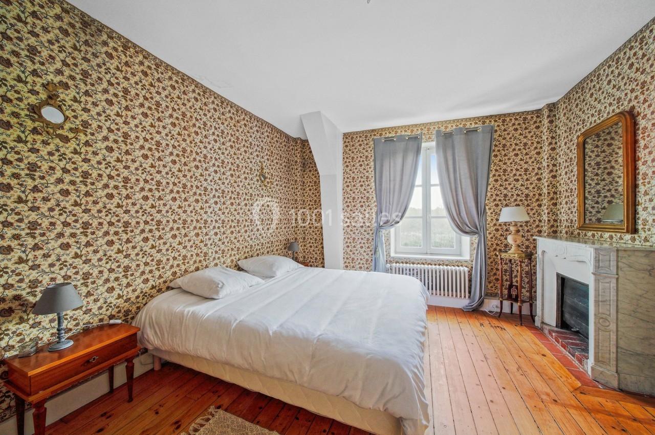 Chambre avec lit double, murs tapissés de motifs floraux, parquet en bois, cheminée en marbre et fenêtre avec rideaux gris.