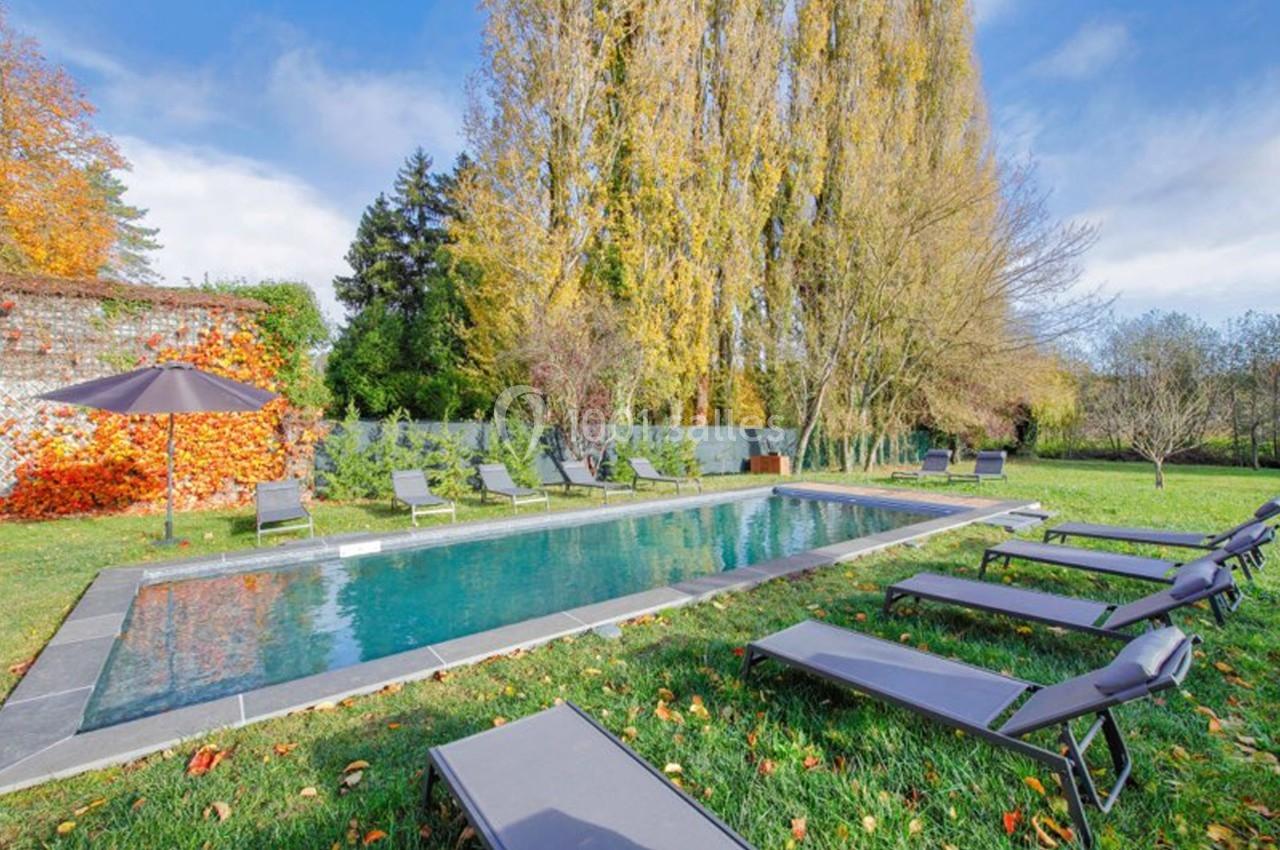 Piscine extérieure entourée de transats, pelouse et arbres aux feuilles d'automne sous un ciel dégagé.