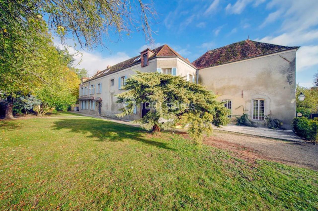 Grande maison ancienne avec toit en tuiles, entourée d'un jardin arboré sous un ciel partiellement nuageux.