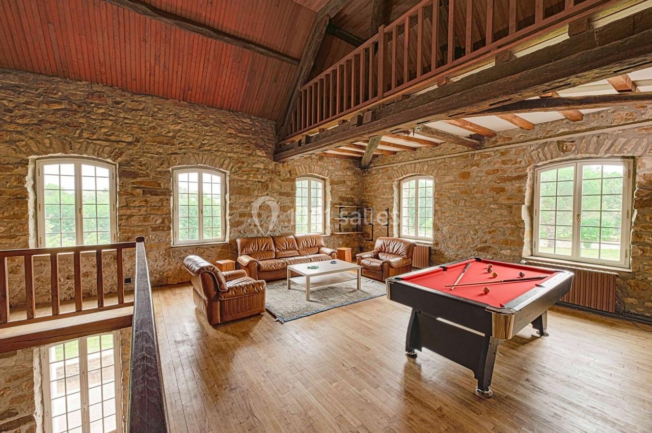 Salle avec murs en pierre, billard rouge, canapés en cuir, table basse et mezzanine en bois.