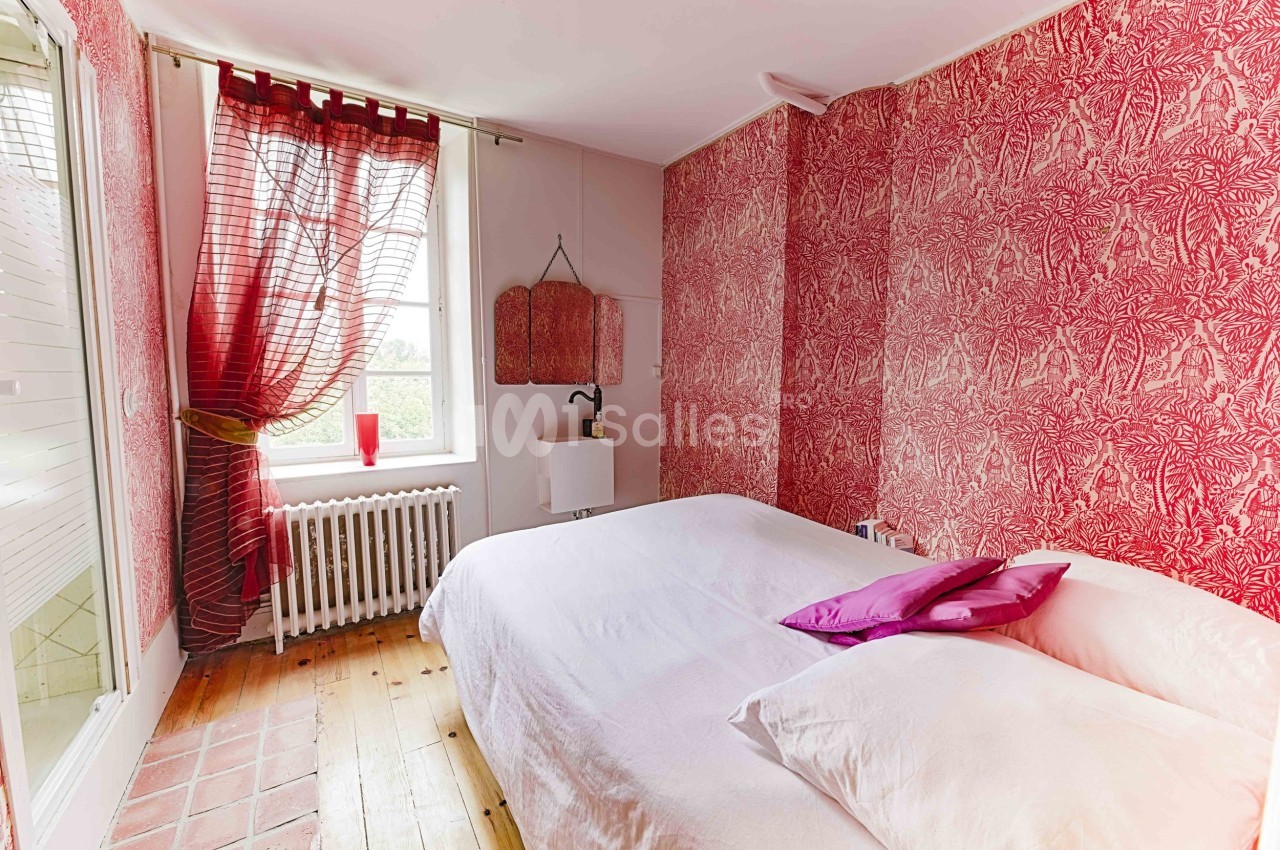 Chambre avec lit double, murs rouges à motifs, parquet en bois et fenêtre avec rideaux laissant passer la lumière.