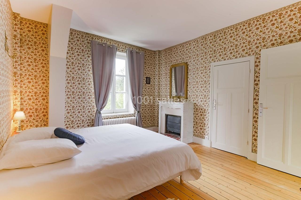 Chambre lumineuse avec lit double, parquet en bois, cheminée décorative et murs tapissés de motifs floraux.
