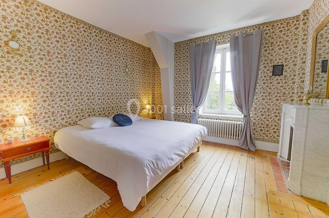 Chambre avec lit double, parquet en bois, papier peint à motifs floraux, cheminée blanche et fenêtre avec rideaux gris.