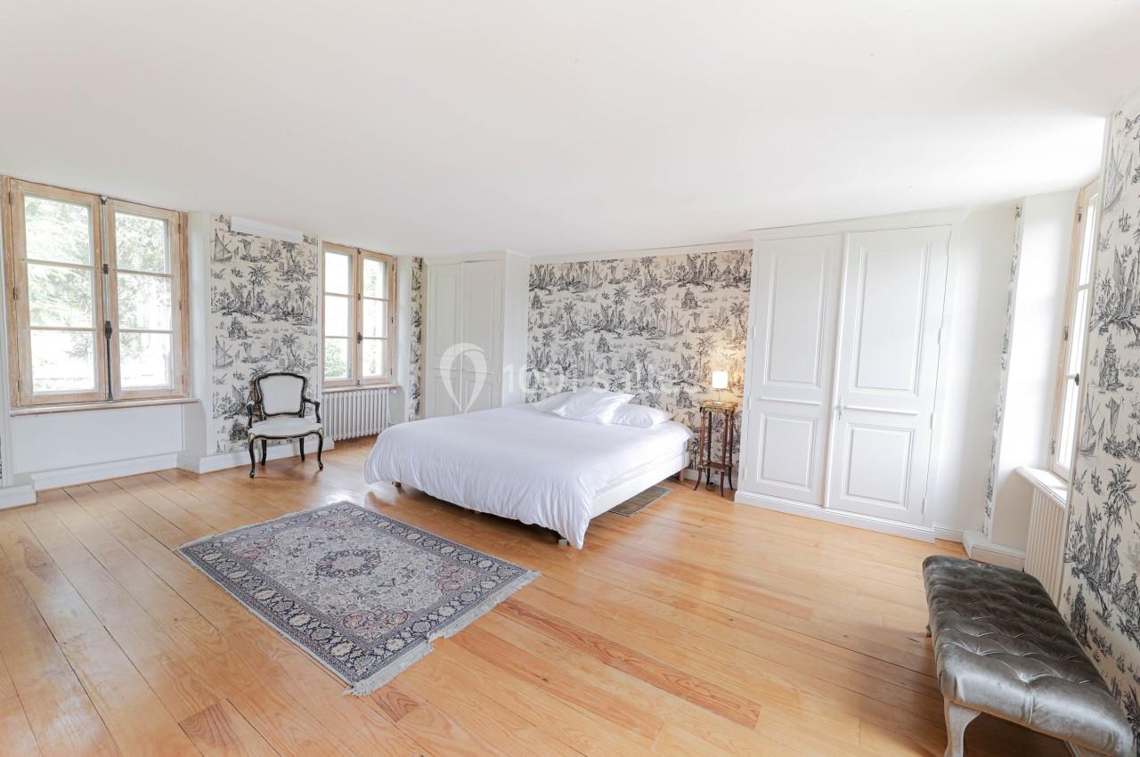 Chambre lumineuse avec lit double, parquet en bois, tapis, murs tapissés et grandes fenêtres donnant sur l'extérieur.