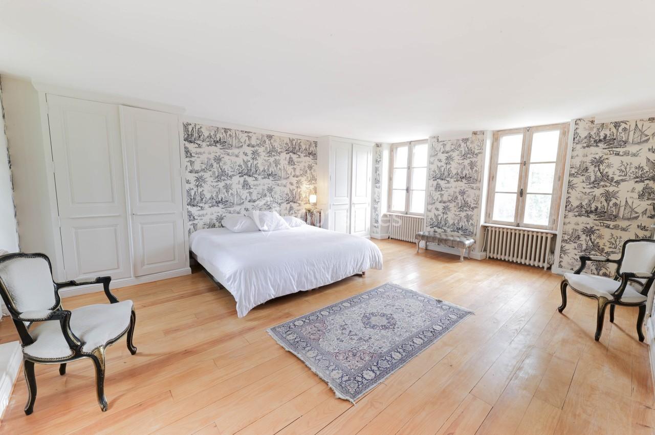 Chambre lumineuse avec lit double, parquet en bois, murs ornés de papier peint à motifs et fenêtres à double battant.