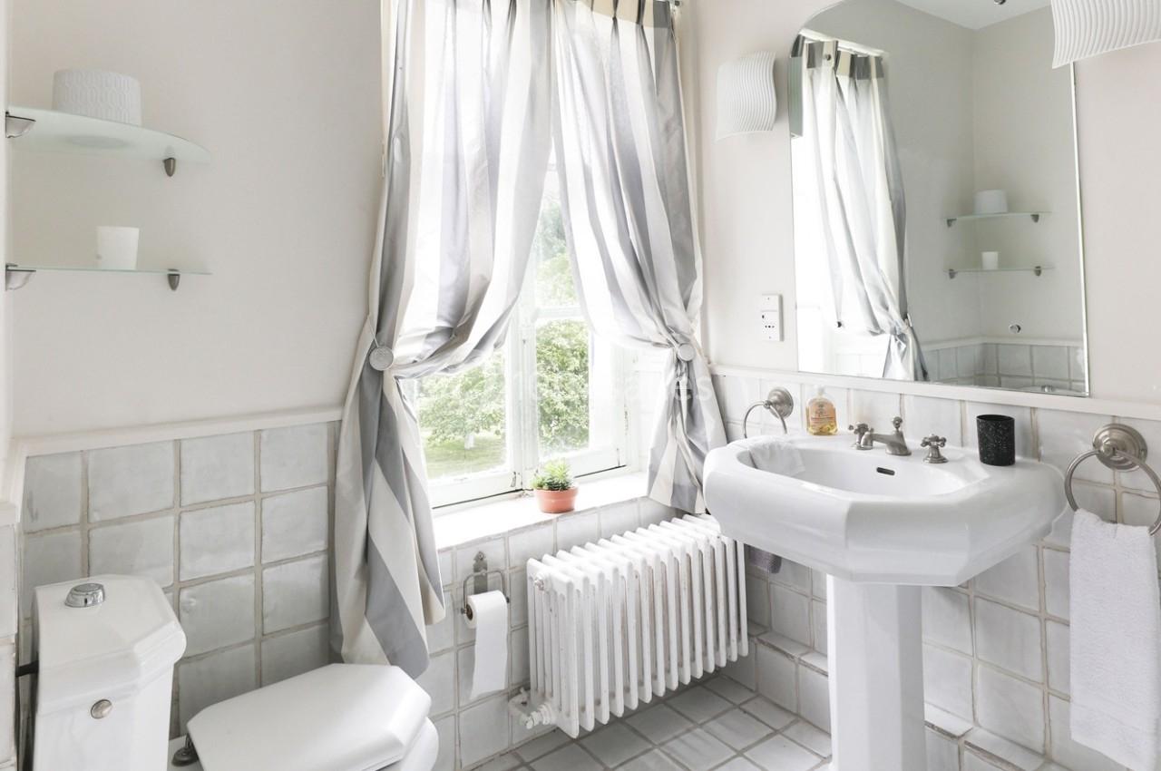 Salle de bain lumineuse avec lavabo, toilettes, miroir, rideaux rayés et vue sur un jardin par une fenêtre.