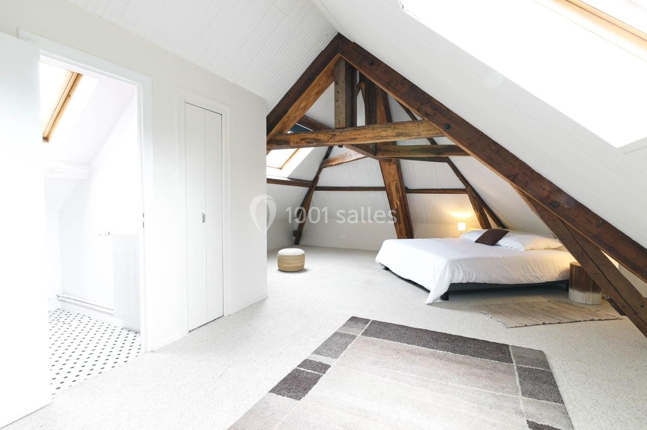 Chambre mansardée lumineuse avec poutres apparentes, lit double, tapis et accès à une salle de bain attenante.