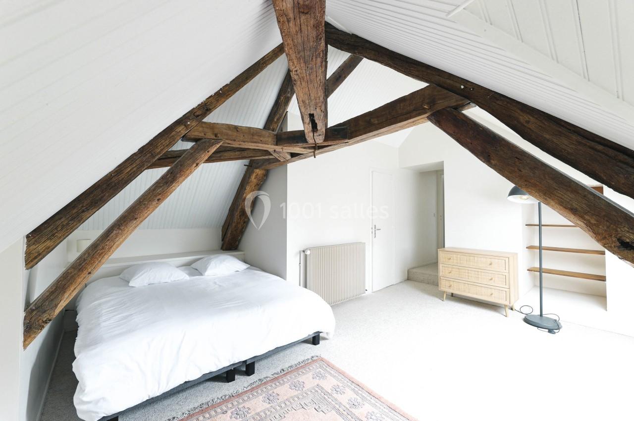 Chambre mansardée lumineuse avec poutres apparentes, lit double, commode en bois, étagères et lampe sur pied.