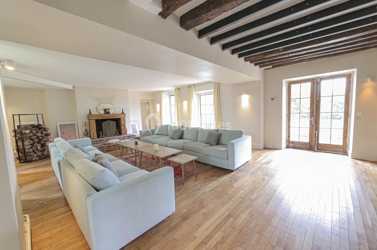 Salon spacieux avec parquet en bois, canapés clairs, cheminée, poutres apparentes et grandes portes vitrées.