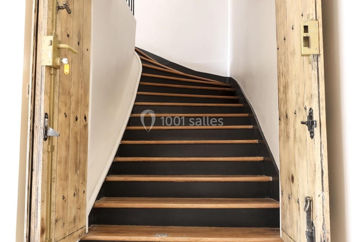 Escalier en bois sombre avec des contremarches claires, encadré par des portes en bois brut ouvertes.