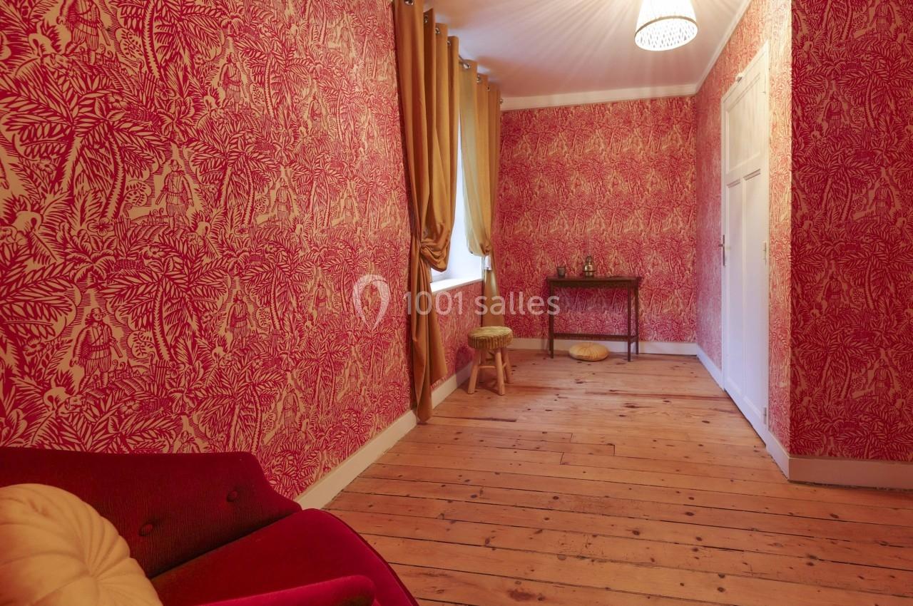 Pièce avec parquet en bois, murs recouverts de papier peint rouge à motifs, rideaux dorés et mobilier minimaliste.