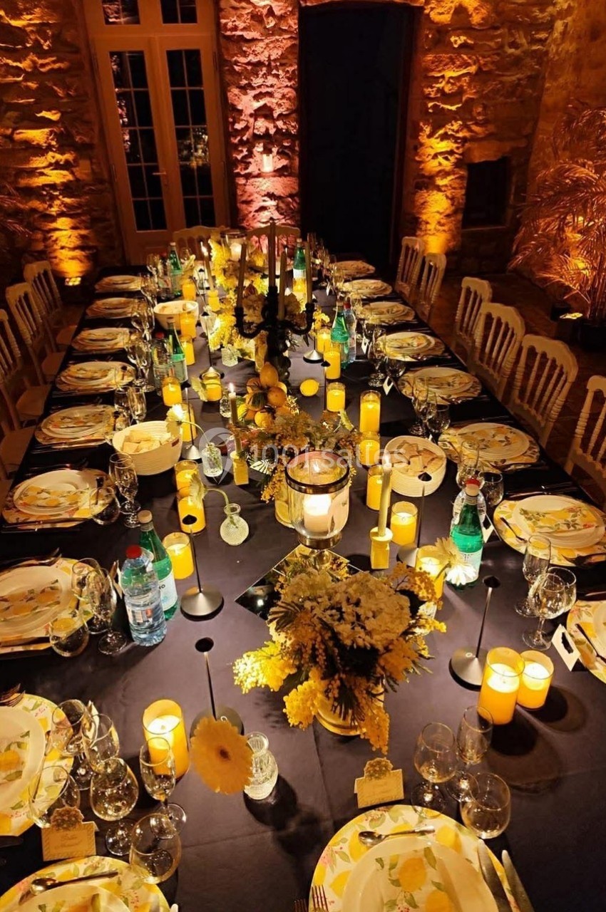 Table élégamment dressée avec des bougies, vaisselle décorée de motifs jaunes et fleurs, dans une salle en pierre éclairée.