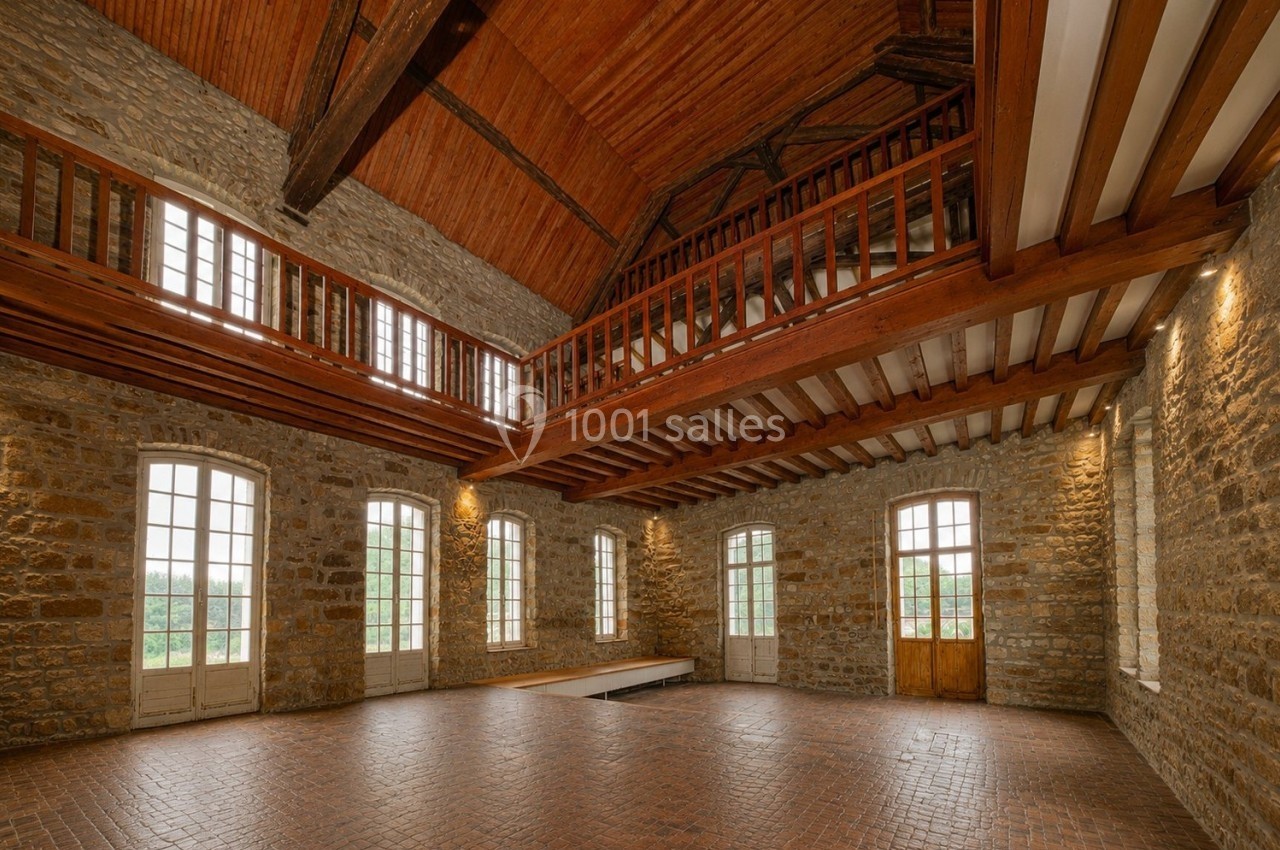 Salle spacieuse avec murs en pierre, plafond en bois, mezzanine et grandes fenêtres laissant entrer la lumière naturelle.