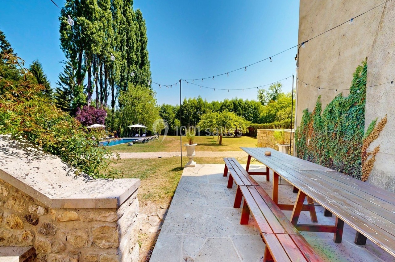 Vue d'une terrasse avec table en bois, jardin arboré, piscine et guirlandes lumineuses sous un ciel bleu.