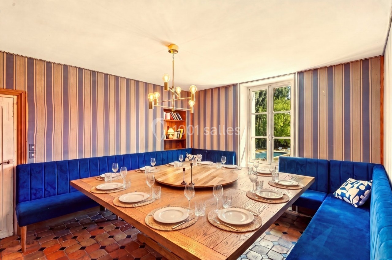 Salle à manger avec table en bois dressée, banquettes bleues, lustre moderne et fenêtre donnant sur un jardin.