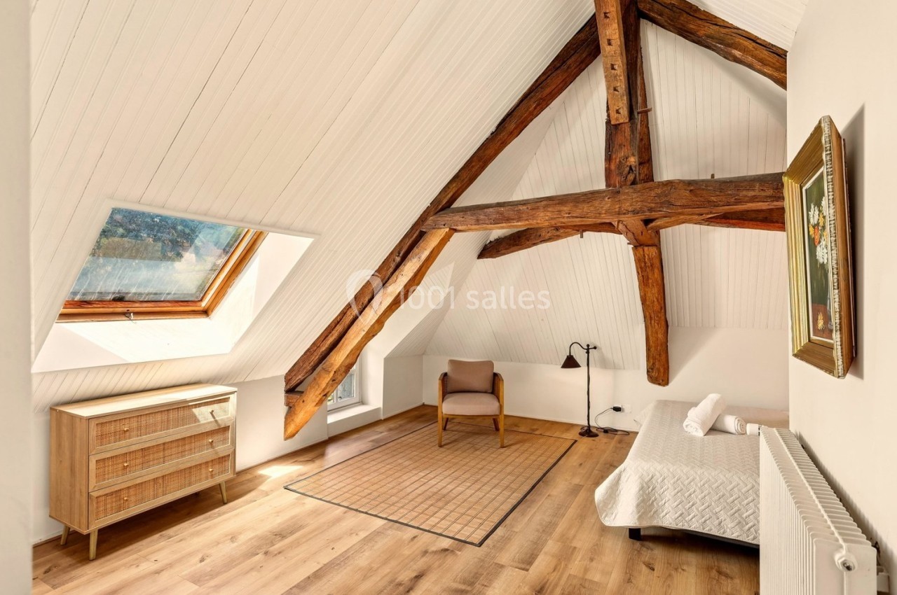 Chambre mansardée lumineuse avec poutres apparentes, lit simple, fauteuil, commode en bois et fenêtre de toit.
