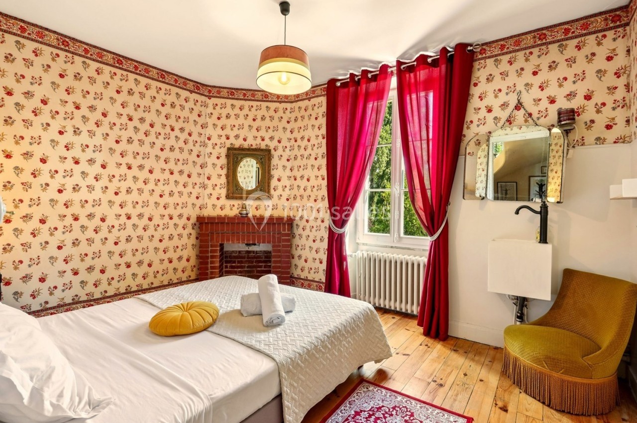 Chambre décorée de motifs floraux avec lit, fauteuil jaune, cheminée, miroir et fenêtre encadrée de rideaux rouges.