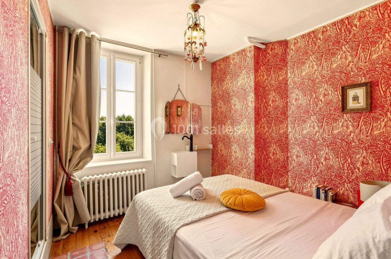 Chambre lumineuse avec murs rouges décoratifs, lit double, coussin jaune, fenêtre ouverte sur un paysage verdoyant.