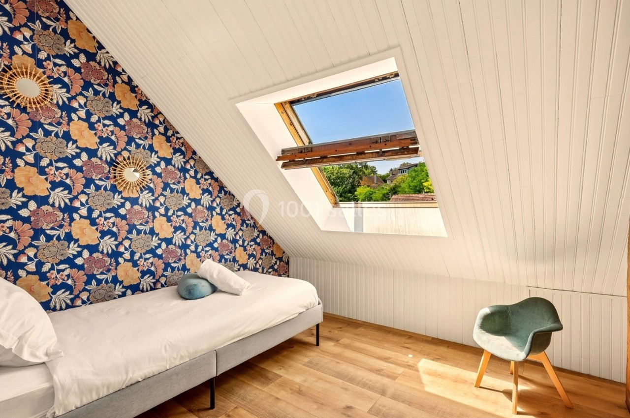 Chambre mansardée avec lit simple, mur floral, fenêtre de toit ouverte et chaise design sur parquet clair.