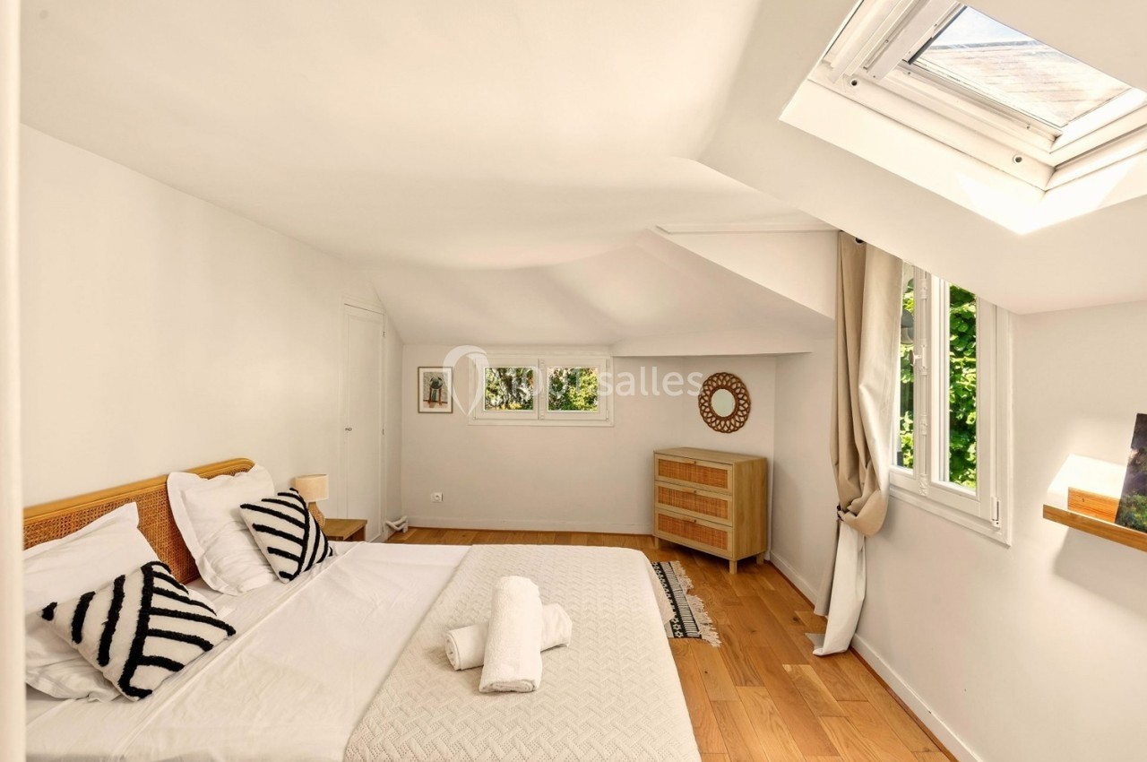 Chambre lumineuse avec lit double, parquet en bois, commode, fenêtres et puits de lumière donnant sur un extérieur verdoyant.