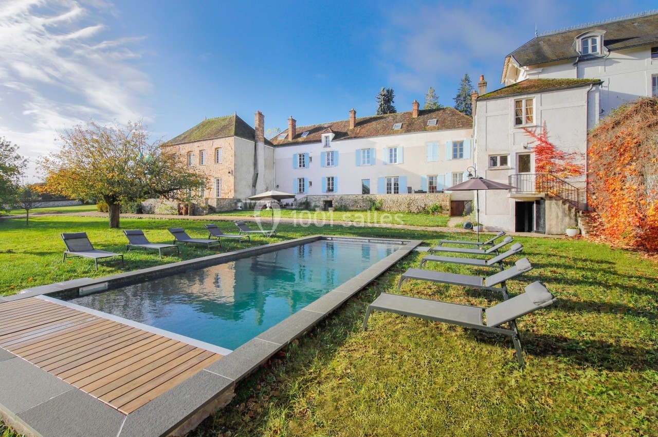 Piscine extérieure entourée de transats sur une pelouse, devant un grand bâtiment ancien sous un ciel dégagé.