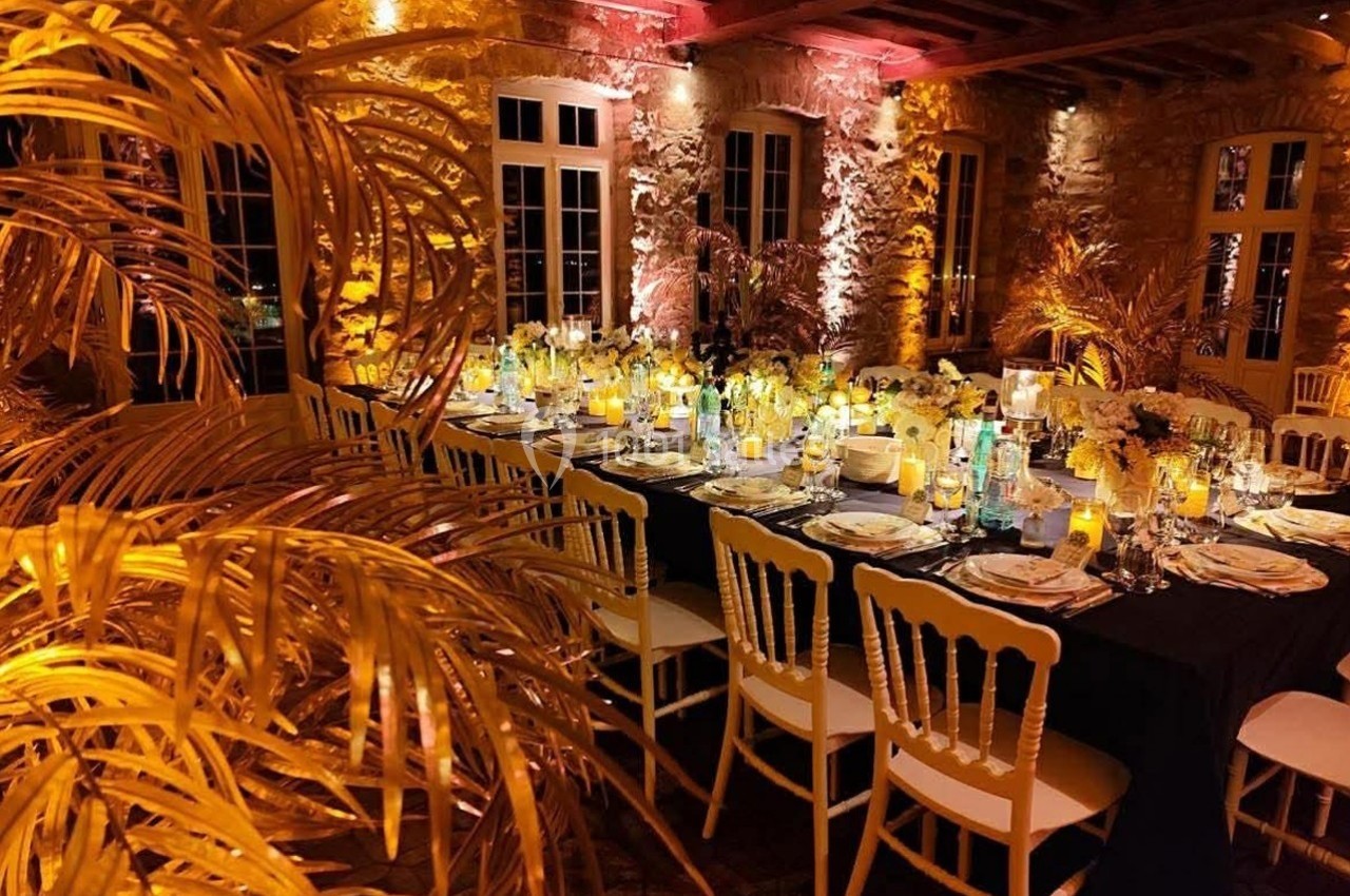 Salle de réception décorée avec des tables dressées, éclairage chaleureux et plantes dorées en premier plan.