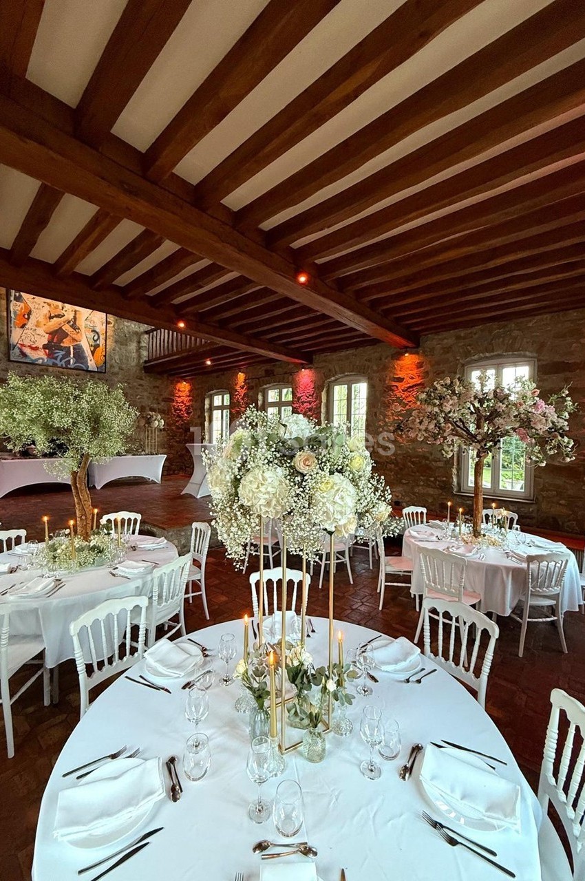 Salle de réception décorée avec des tables rondes, nappes blanches, chandeliers dorés et compositions florales élégantes.