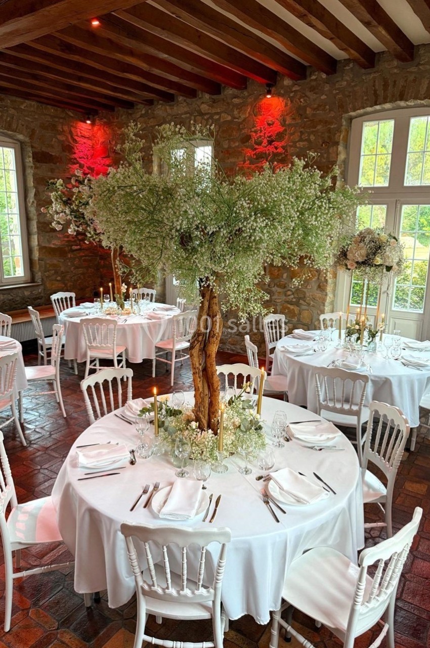 Salle de réception avec tables rondes décorées de nappes blanches, chaises en bois et centres de table fleuris.