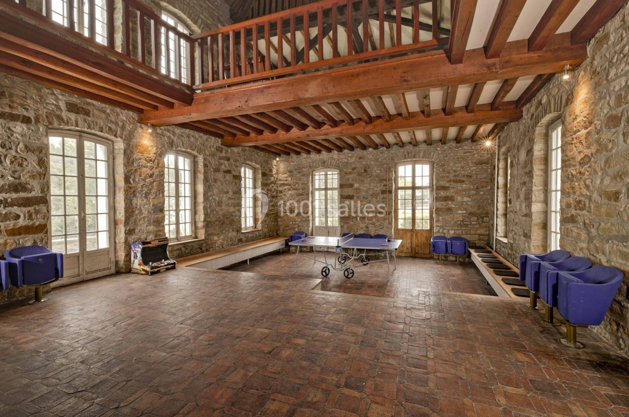 Salle spacieuse avec murs en pierre, plafond en poutres apparentes, table de ping-pong et fauteuils bleus.