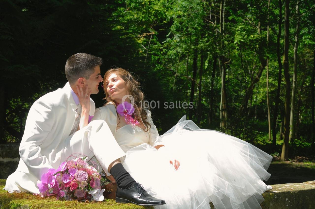Un couple en tenue de mariage assis sur un muret, entouré de verdure, avec un bouquet de fleurs posé à côté.