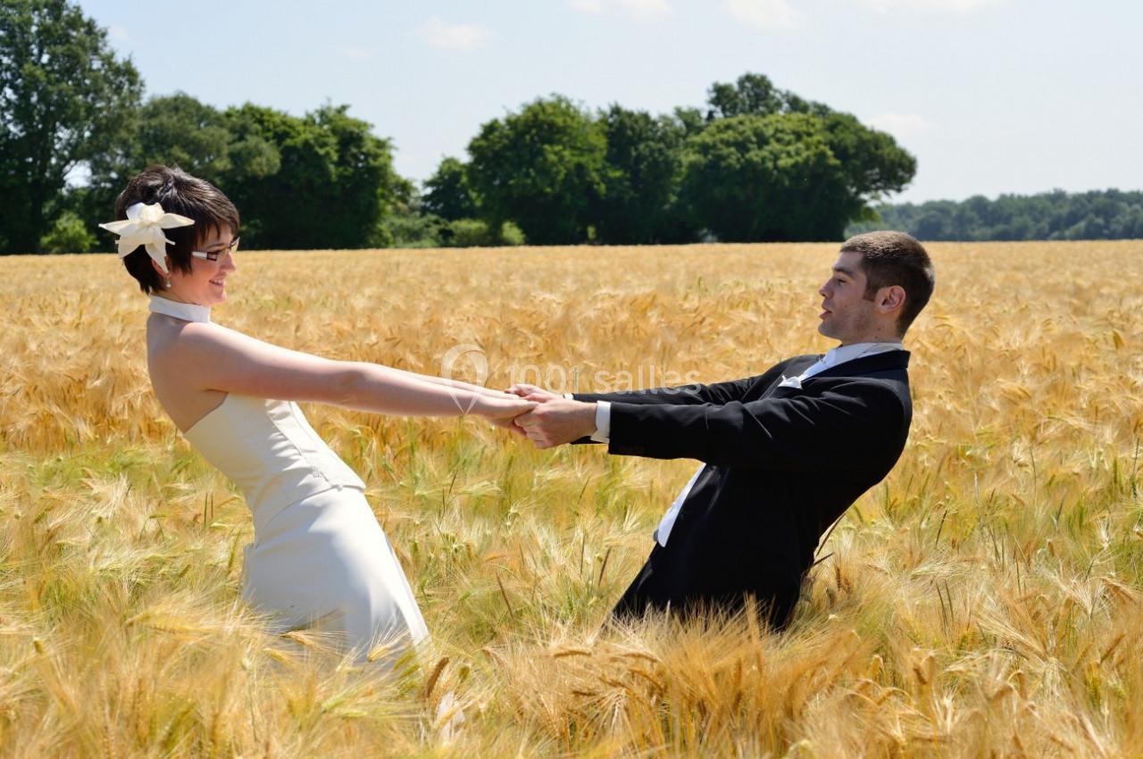 Un couple en tenue de mariage se tient par les mains dans un champ de blé sous un ciel bleu.