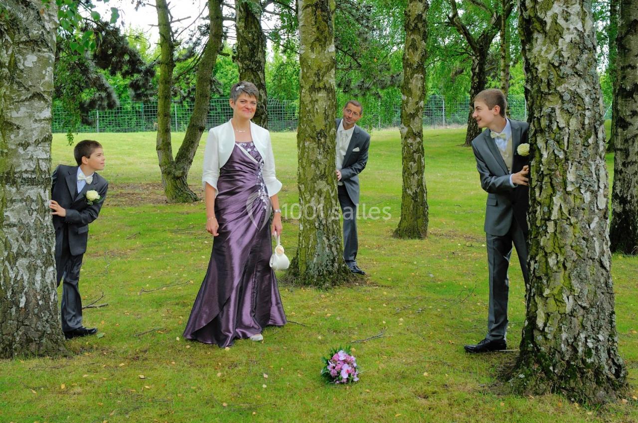 Une femme en robe de soirée pose dans un parc, entourée de trois hommes en costume se cachant derrière des arbres.