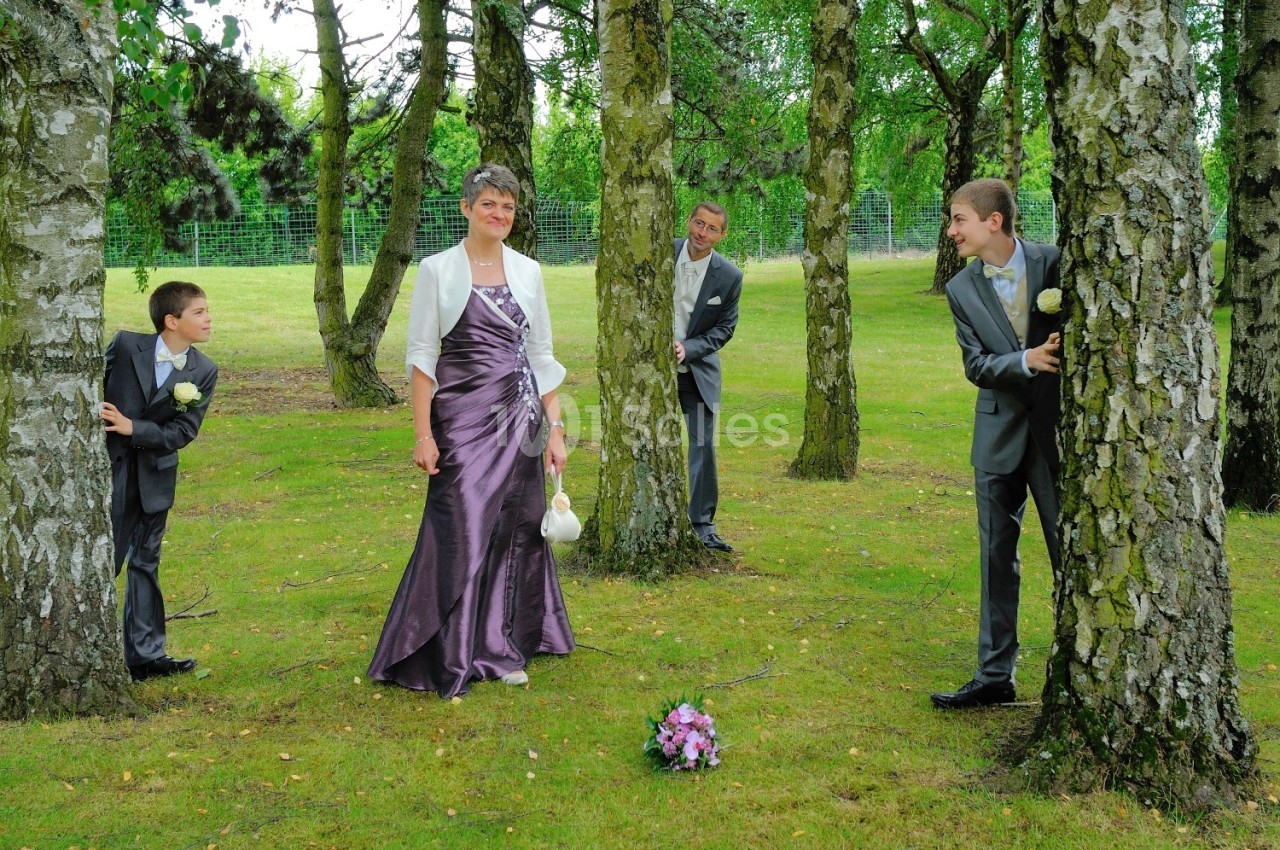 Une femme en robe de soirée pose dans un parc, entourée de trois hommes en costume se cachant derrière des arbres.