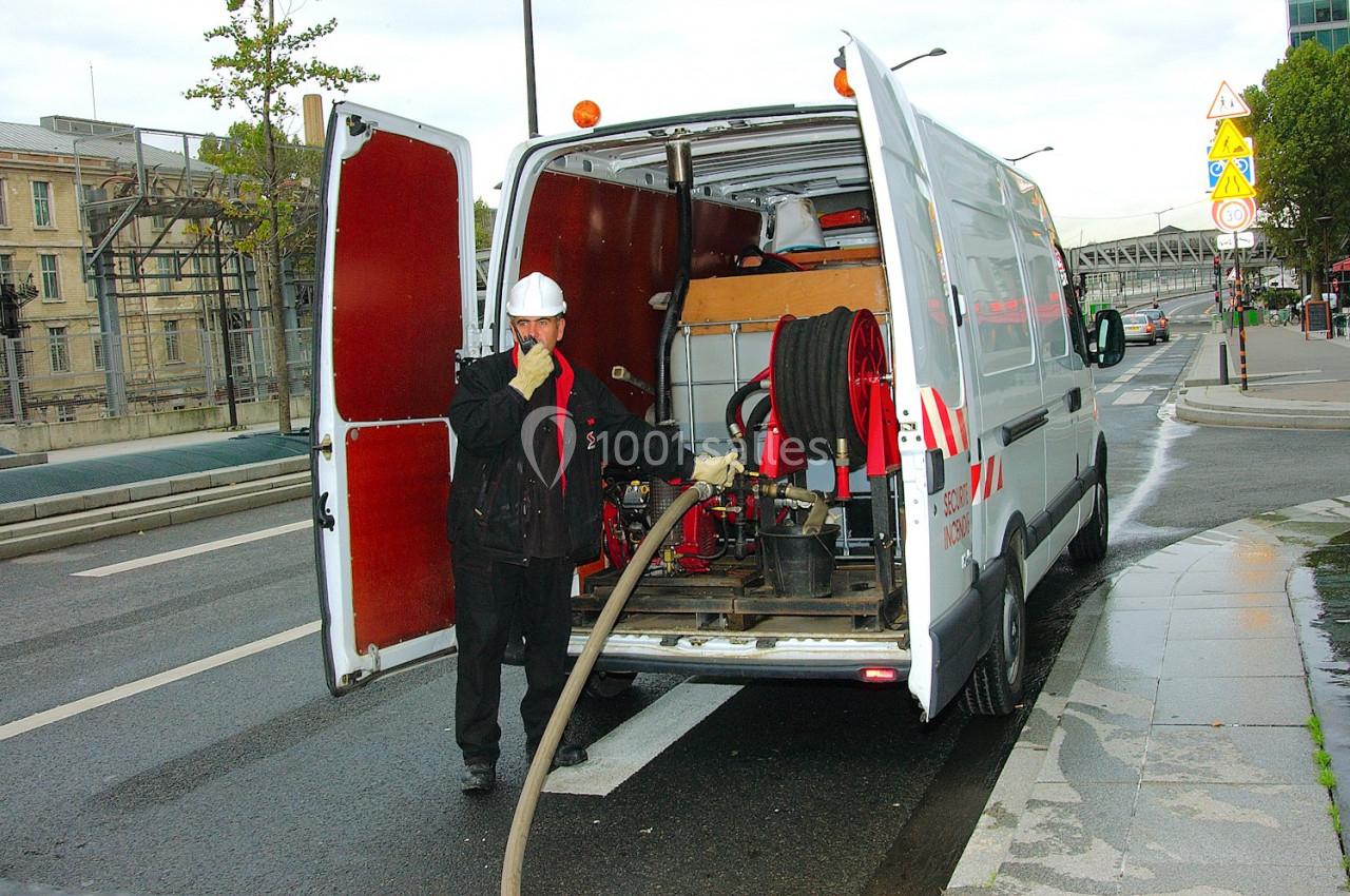 Un technicien en tenue de sécurité utilise un tuyau relié à un fourgon équipé de matériel technique sur une route.