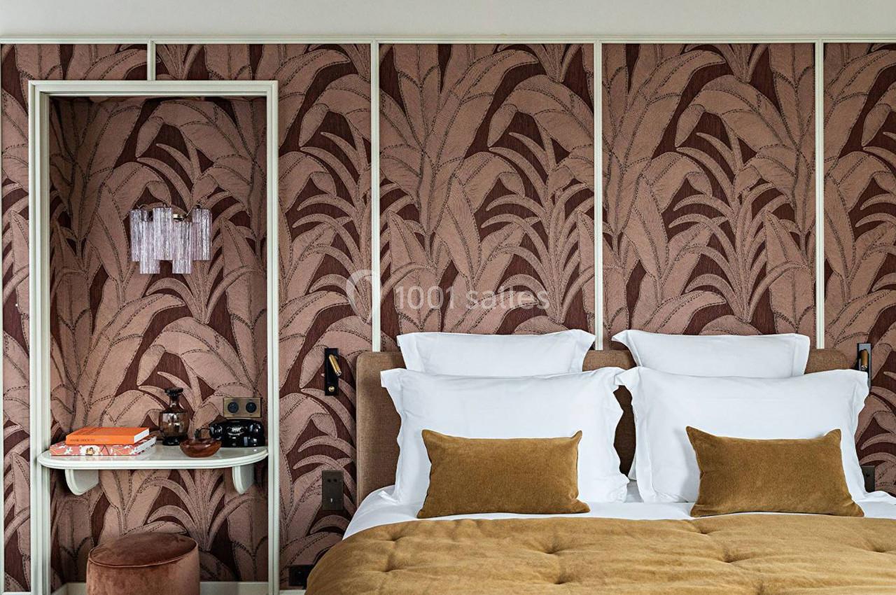 Chambre avec lit double, coussins marron, murs décorés de motifs végétaux et petite étagère avec objets.