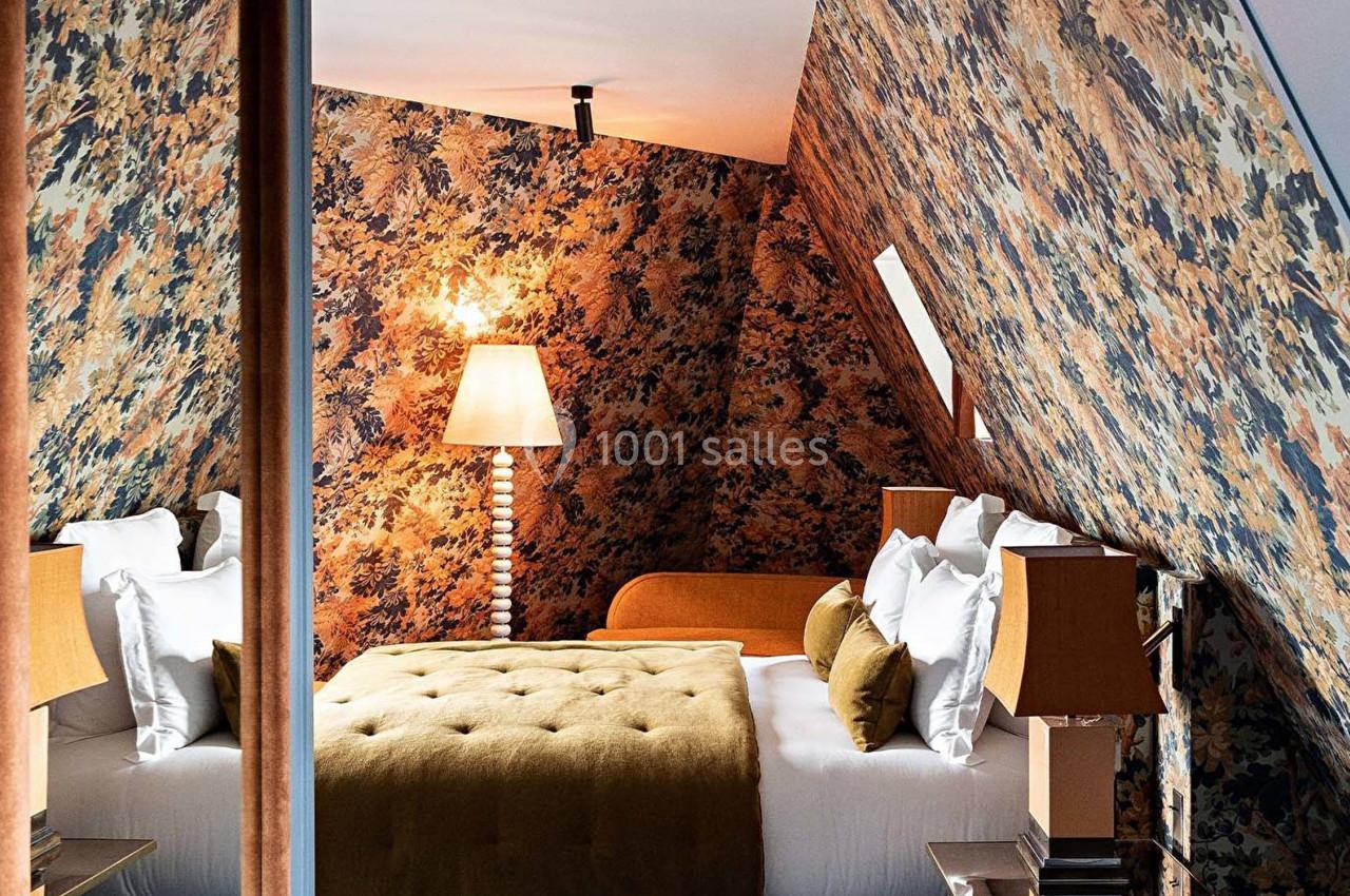 Chambre mansardée avec papier peint à motifs floraux, lit double, lampes de chevet et éclairage chaleureux.