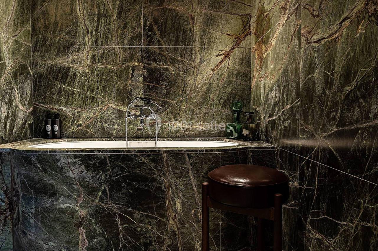 Baignoire encastrée entourée de murs et sol en marbre vert foncé, avec un tabouret en cuir marron au premier plan.