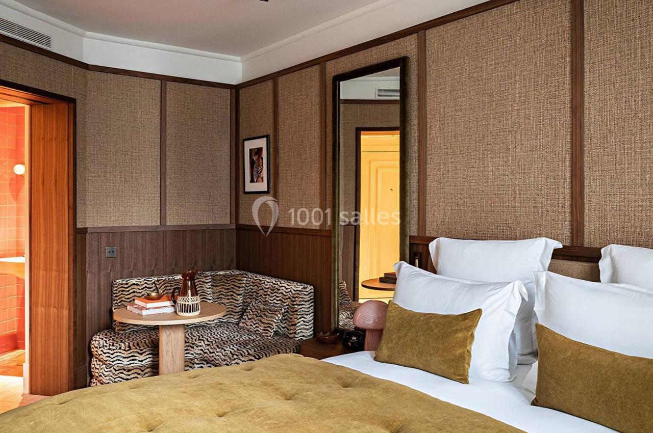 Chambre d'hôtel avec lit double, coussins jaunes, canapé imprimé, table basse et vue partielle sur une salle de bain.