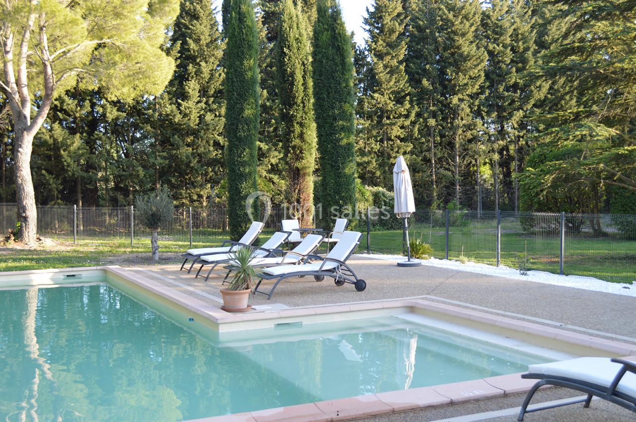 Piscine extérieure entourée de chaises longues, parasol et arbres, dans un cadre naturel verdoyant.