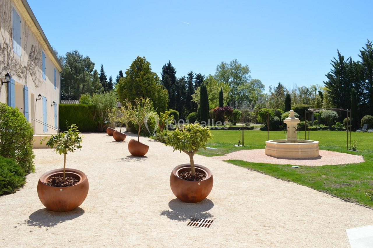 Allée bordée de pots en terre cuite avec des arbustes, menant à une fontaine dans un jardin verdoyant.
