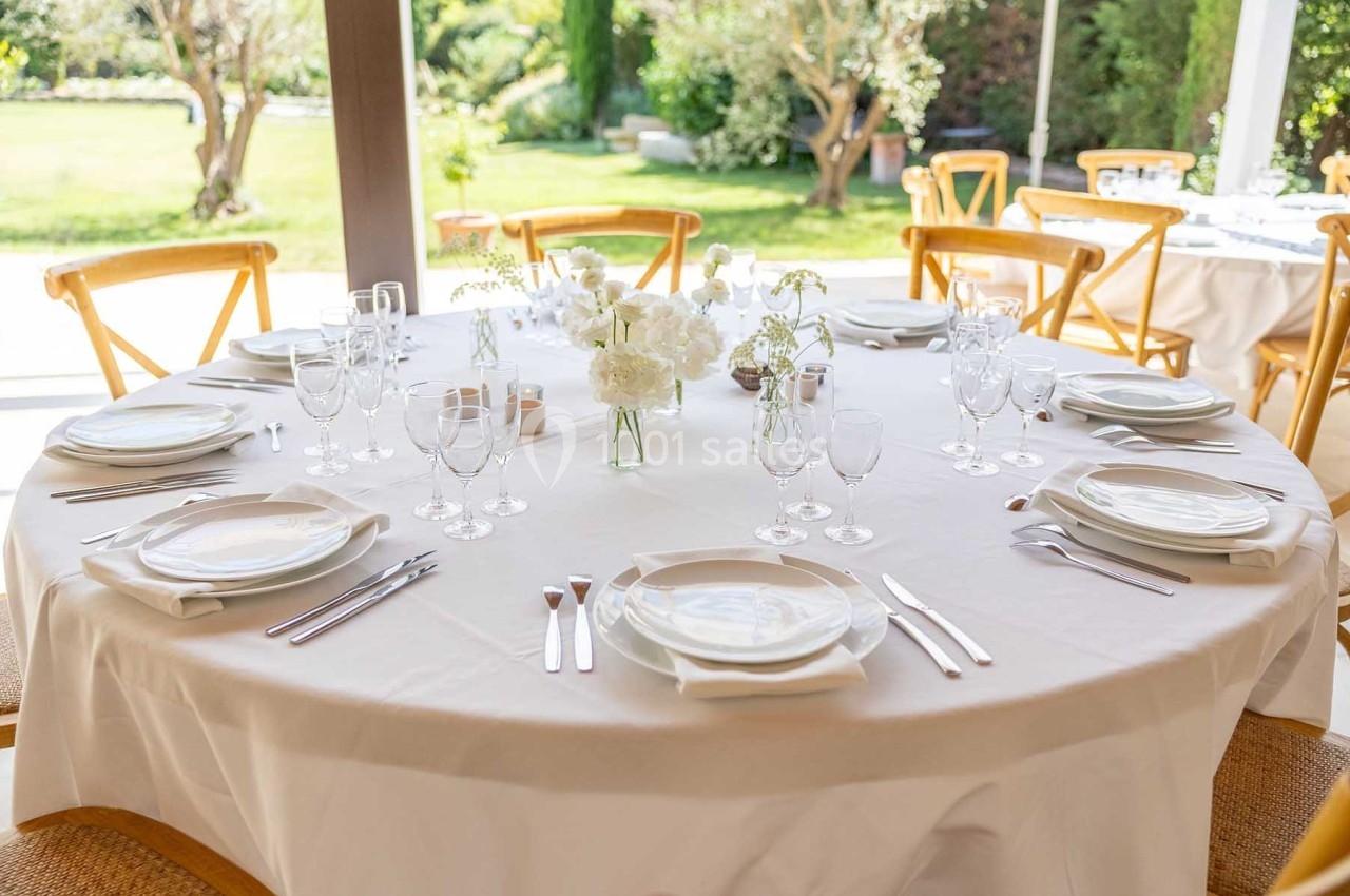 Table ronde élégamment dressée avec nappes blanches, vaisselle, verres et centre de table floral, en extérieur.