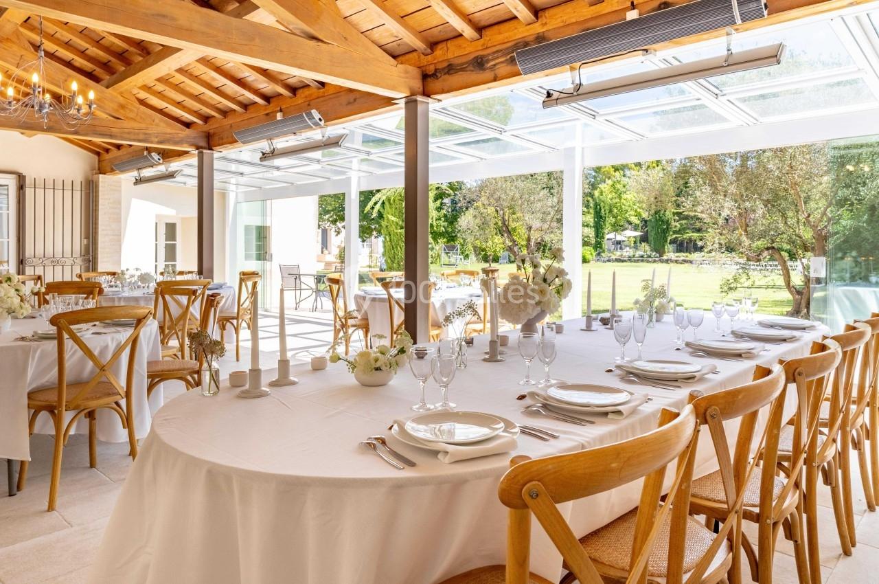 Salle lumineuse avec tables rondes dressées, chaises en bois, grandes baies vitrées et vue sur un jardin verdoyant.
