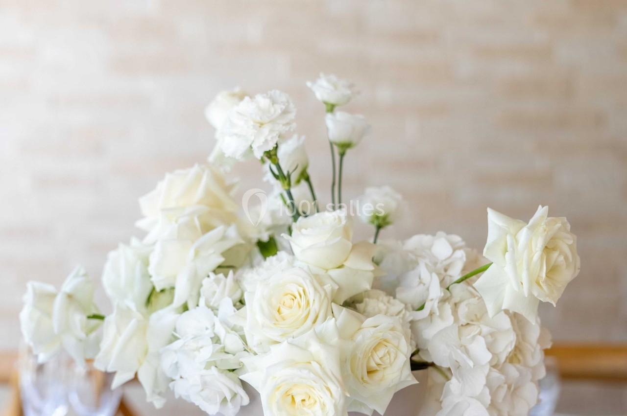Arrangement de fleurs blanches, incluant des roses et des hortensias, dans un vase beige sur une table blanche.