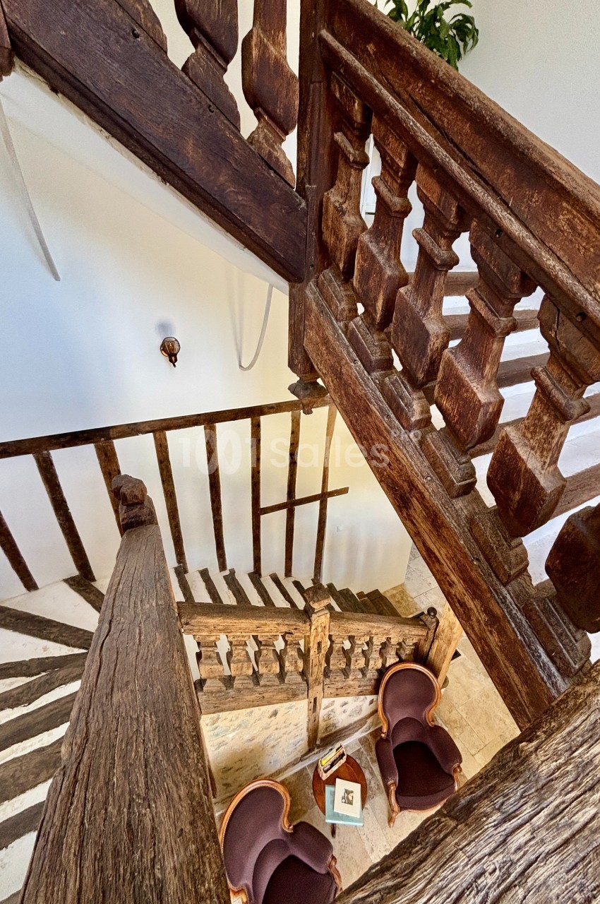 Escalier en bois ancien avec balustrades sculptées, vu en plongée, entouré de murs blancs et de fauteuils en bas.