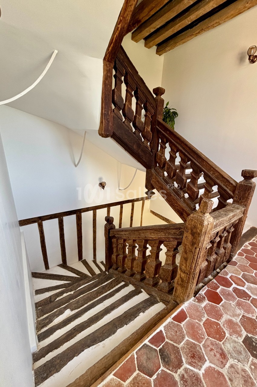 Escalier en bois ancien avec rambardes sculptées, murs blancs et sol en carreaux de terre cuite.