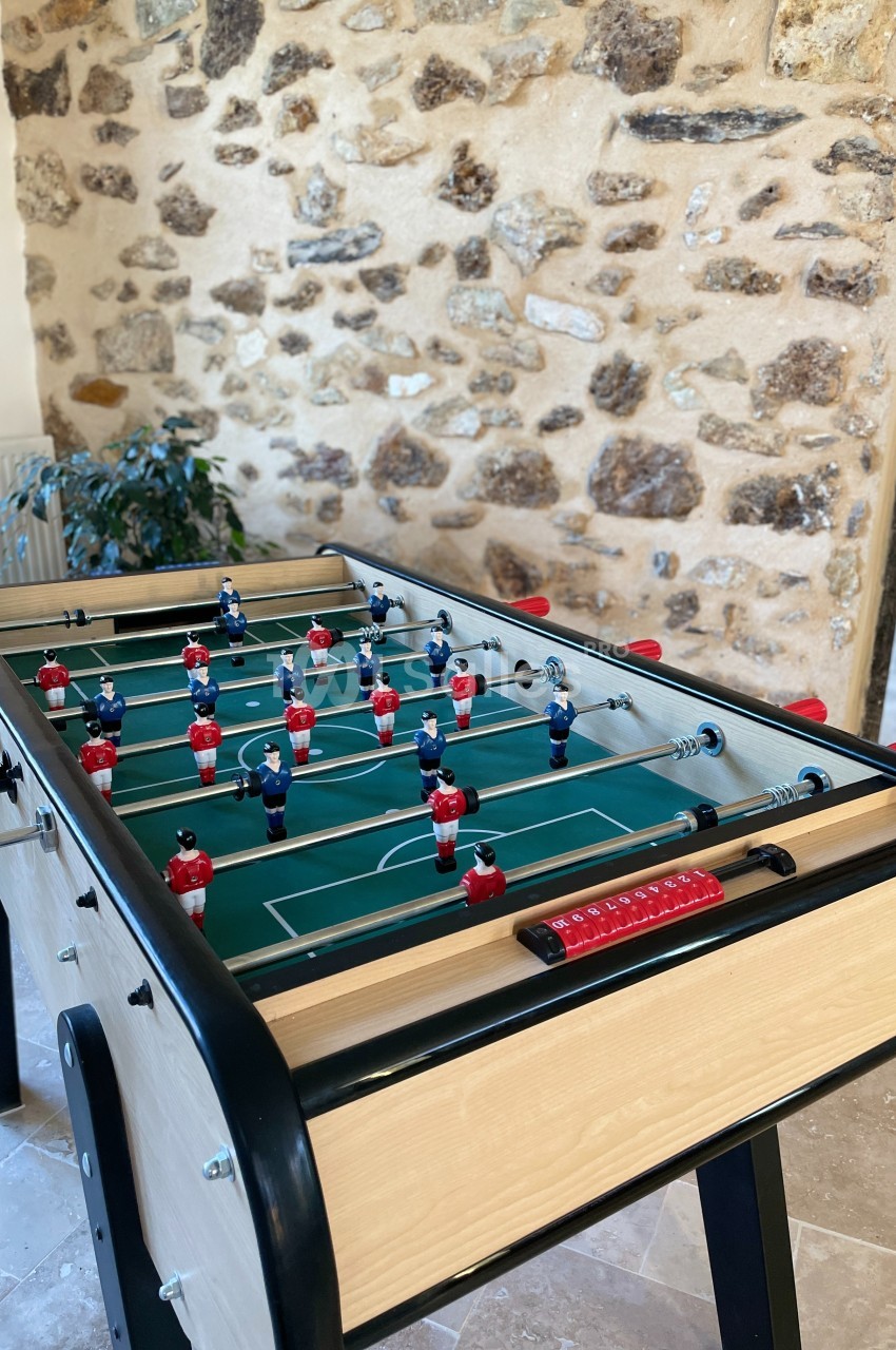 Table de baby-foot en bois clair avec figurines rouges et bleues, placée dans une pièce aux murs en pierre apparente.