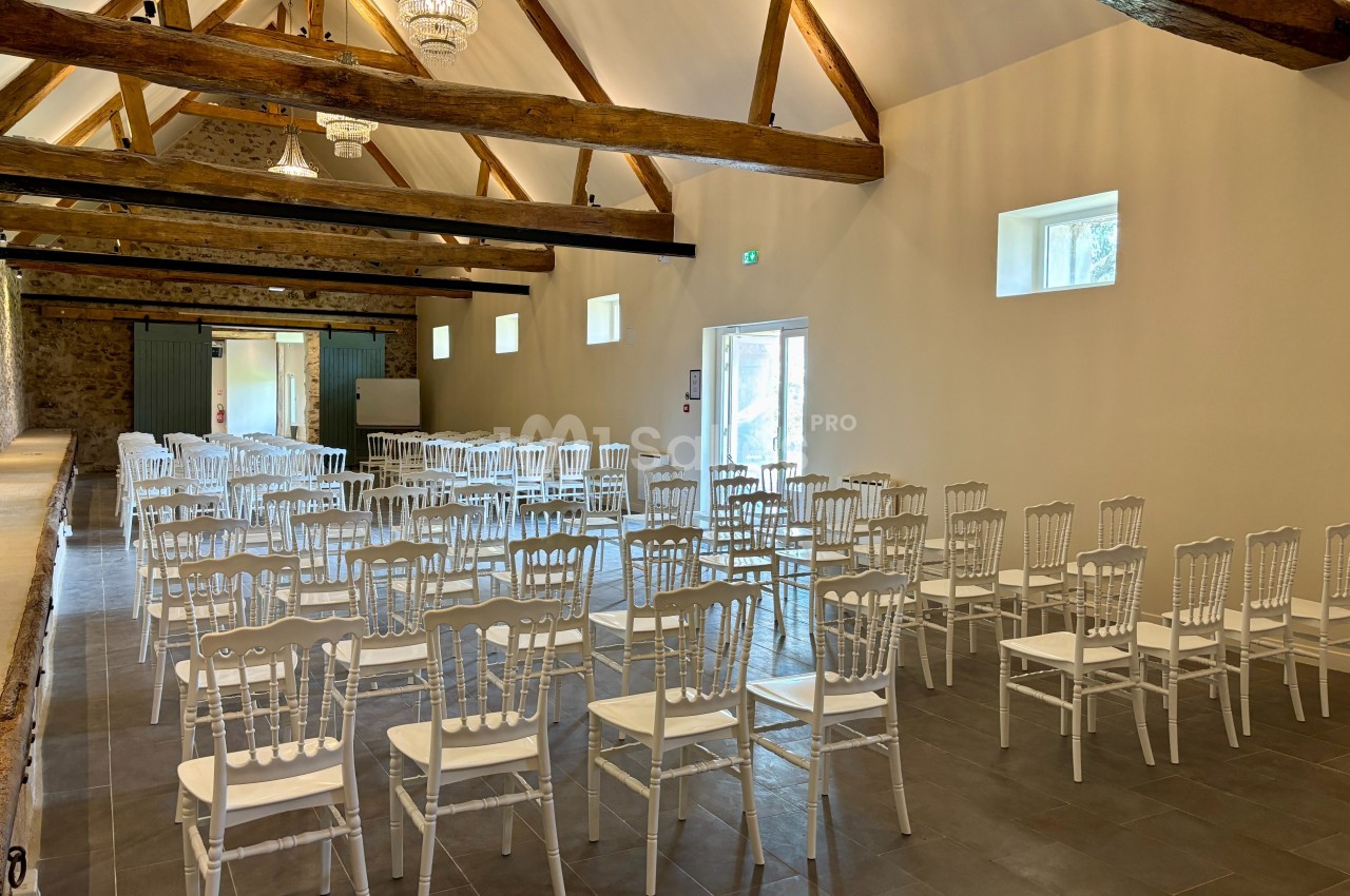 Salle lumineuse avec des chaises blanches alignées, poutres apparentes au plafond et murs en pierre.